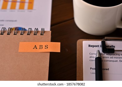 There's A Cup Of Coffee,dummy Document Paper And A Notebook With A Sticky Note On It That Says ABS Written On It. It Was An Abbreviation For The Financial Term Asset Backed Security.