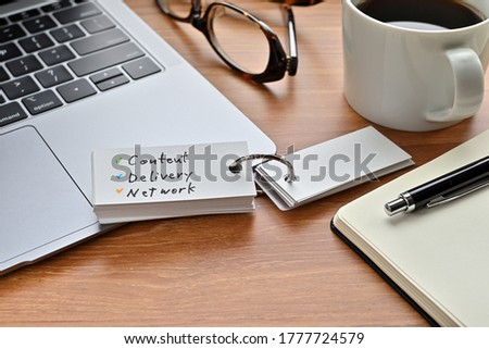 There's a laptop computer, a notebook, a pen and an open vocabulary book on the desk. The word Content Delivery Network is written on it.
