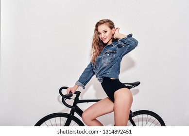 There's no fear when you're having fun. Seductive young lady in bodysuit and denim jacket looking at camera. She is going to ride a bike, isolated over white background
