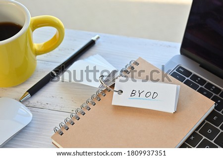 There's a word book on a desk with a cup of coffee and laptop pc. The word BYOD is written in it. It's an abbreviation for Bring Your Own Device.