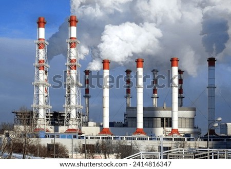 Thermoelectric power station building with many high red and white industrial pipes with dense smoke under winter sky on sunny day front view