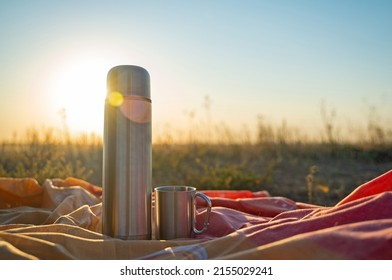 Thermos and camping cup on a bedspread against the backdrop of sunset or sunrise