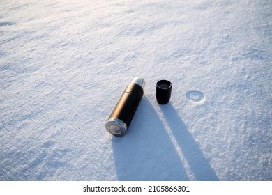 The Thermos Lies On The Snow. The Shadow Of The Dishes Falls On The Snow. A Hot Drink Of Coffee Is Poured Into A Thermal Mug. High Quality Photo