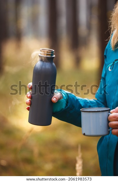 Thermos with\
steaming hot drink. Refreshment during hiking. Woman holding travel\
mug and insulated drink\
container