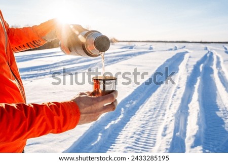 A thermos with tea on the background of snow, the glare of the sun shines into the camera, hot tea pours from a bottle into a cup, winter recreation in nature. High quality photo