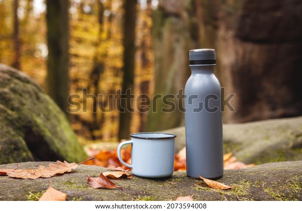 Thermos and travel mug in\
autumn forest. Insulated drink container on rock. Refreshment\
during hiking