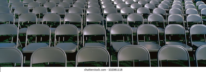 These Are Empty, Grey Folding Chairs Awaiting A Crowd To Attend A Graduation Ceremony. They Are Neatly Set Up In Rows Side By Side.