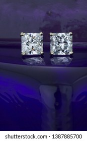 These square cut diamonds are mounted in four prong platinum earring post. On a reflective blue  background.
