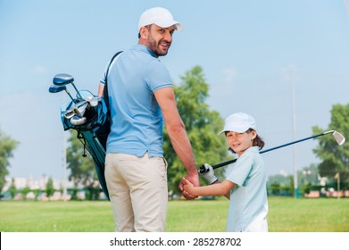 They are the big golf fans. Happy young man and his son holding hands and looking at camera while walking on the golf course