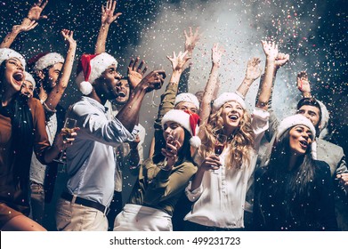 They Love Christmas. Group Of Beautiful Young People In Santa Hats Throwing Colorful Confetti And Looking Happy