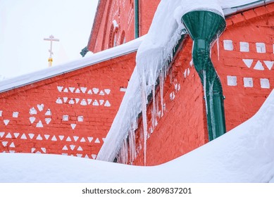 A thick, cascading layer of snow and long icicles hangs from the roof edge beside a green drainpipe, set against a red brick church with white geometric patterns