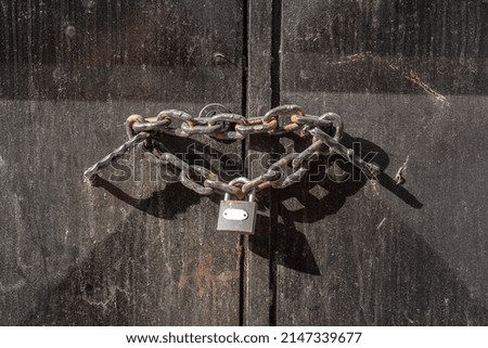 Thick chain with closed lock is threaded through the iron handles on iron door. Black dirty door.