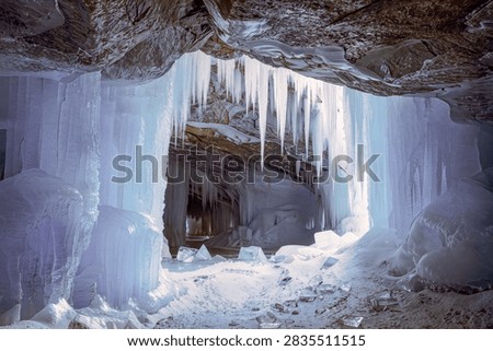 Thick columns and needle sharp icicles frame the mouth of a frozen sea cave where daylight pushes through the ice and turns the whole chamber pale blue. Slabs of broken ice litter the snow covered floor beneath the hanging formations.