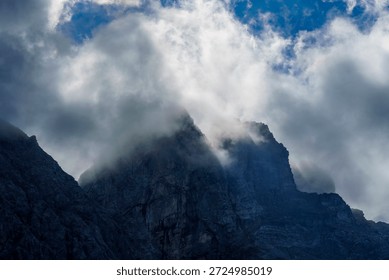 Thick, dramatic clouds swirl around a dark, jagged mountain peak, partially obscuring the rocky summit under a dynamic sky.