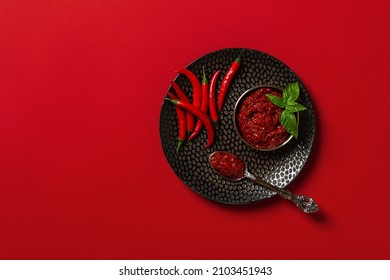 Thick hot chili sauce in a bowl and heap of fresh red hot chili peppers on a black textured plate over red background. Spices and condiments concept. Copy space. Flat lay. Top view.
