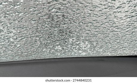 Thick layer of patterned ice covering a car windshield viewed from the dark interior dashboard. Dangerous winter driving conditions and freezing rain