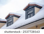 Thick layers of snow blanket the cozy rooftops, accentuated by dark dormer windows, creating a picturesque winter scene under a cloudy sky.