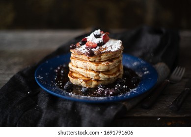 Thick pancakes are stacked on a plate covered in blueberries, strawberries and syrup ready for breakfast or brunch