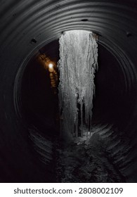A thick pillar of ice comes down from the ceiling inside a corrugated metal culvert. The wet, icy floor leads to a bright sunrise at the end of the tunnel.