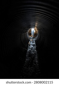 A thick pillar of ice comes down from the ceiling inside a corrugated metal culvert. The wet, icy floor leads to a bright sunrise at the end of the tunnel.