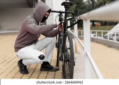 A thief in a balaclava and a hood breaks the lock on a bicycle in the street during the day. Closed face and hacking. Stealing bicycles. - Powered by Shutterstock