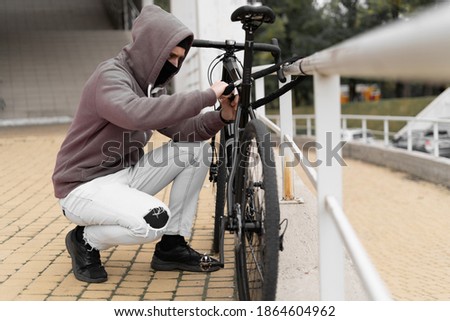 A thief in a balaclava and a hood breaks the lock on a bicycle in the street during the day. Closed face and hacking. Stealing bicycles.