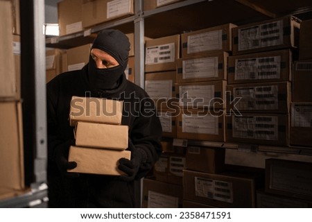 Thief in black balaclava steals with a parcel in a warehouse at the night. Concept of security problems in storehouses and stores. Copy space