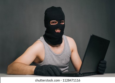 A thief in a black mask stole a laptop. A man in a balaclava and a computer in hand on a gray background. The hacker is hacking the computer. Steals information. A young man breaks the law. - Shutterstock ID 1128471116