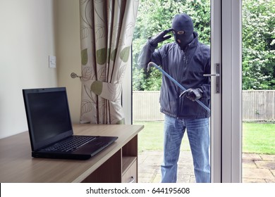 Thief breaking into a house via a patio doors window with a crowbar to steal a laptop computer from an office desk