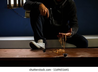 Thief Holding And Looking At The Stolen Gold Necklace With Gun On Old Table. Low Key. Killer Man.

