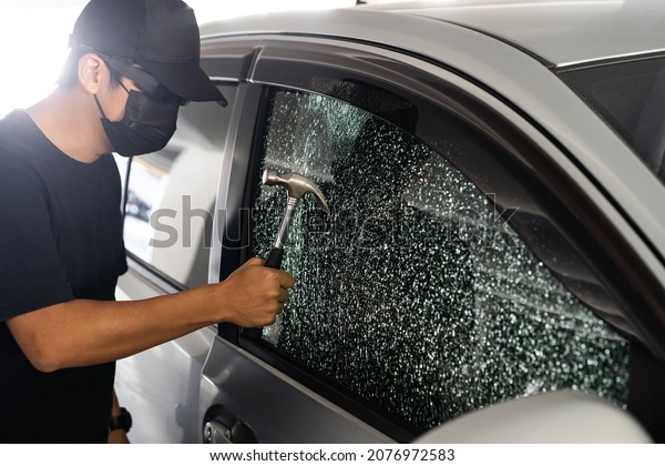 Thief with mask breaking car window\
with hammer on day time. Car thief, car theft concept.\
