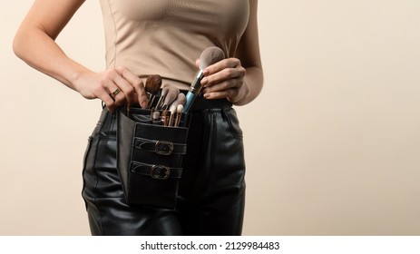 Thigh bag with different brushes on faceless professional makeup artist. Unrecognizable make-up artist with belt bag with tassels or makeup brushes over beige background
