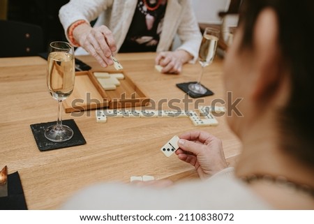 Thin fingers and hands of unrecognizable elderly people possibly women in white jacket playing dominoes in cozy cafe outdoors lying on table with glasses filled with champagne or wine celebrating 