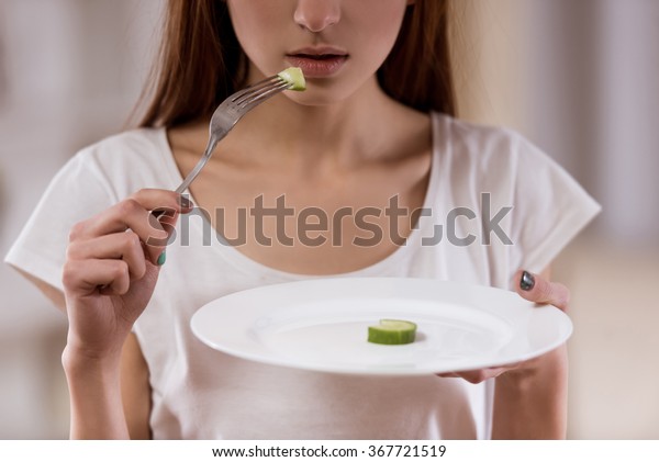 部屋の接写の真ん中に空の皿を持つ薄い女の子が立っている 栄養失調は健康を害する の写真素材 今すぐ編集