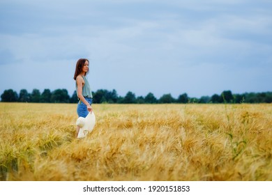A thin girl with a hat in her hand is spinning in the middle of a wheat field against the background of the sky with clouds. Rural summer landscape.