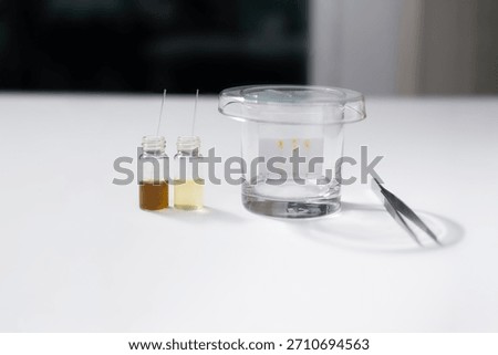 A Thin Layer Chromatography (TLC) plate develops inside a glass tank, separating compounds, accompanied by sample vials and forceps for chemical analysis.