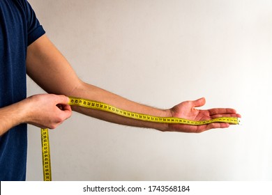 Thin Man Measuring His Left Arm From Fingers To Elbow With A Yellow Tape Measure. White Background. Olive Skin Tone. 