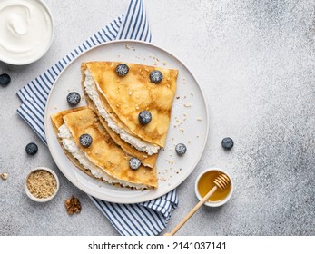 Thin pancakes, russian blini or french crepes with riccotta (curd or cottage cheese), fresh blueberry, honey , sour cream bowl and chopped walnuts. Tasty homemade breakfast. Top view, copy space. 