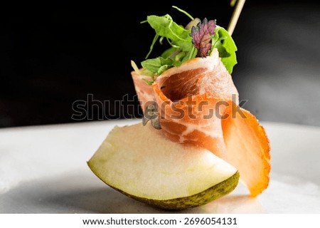 Thin slices of prosciutto ham wrapped around a piece of juicy melon, garnished with fresh greens and served on a white plate. Close-up, selective focus with a dark background