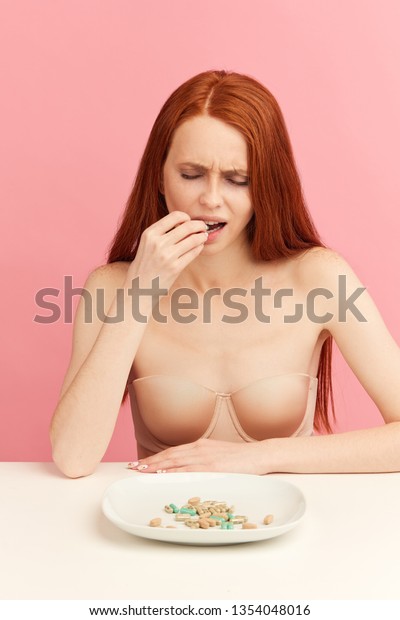 Thin
undressed woman with long ginger hai feeling sick and unhappy
eating anti-obesity pills, obsession with weight loss, addiction to
anti-obesity pills, drugs as easy way to lose
weight