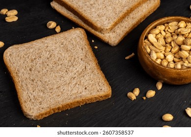 thin wheat bread with bran and soft peanut butter, peanut butter on a piece of bread and roasted peanuts on the table on black slate