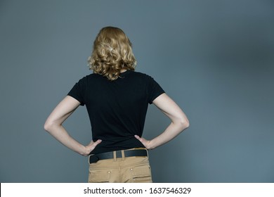 A Thin Young Man With Long Blonde Hair In A Black T-shirt And Beige Jeans, Stands With His Back To The Camera Hands On The Waist