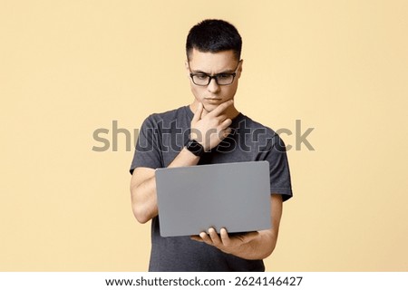 Think about difficult task, online education and work. Pensive busy young man student with glasses presses hand to chin and looks at laptop, isolated on beige background, studio shot, free space