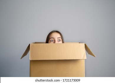 Think outside the box concept. Funny girl with scared face looking out of box at grey background. Getting out of comfort zone. 