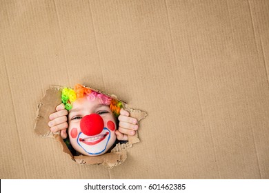Think outside the box. Funny kid clown looking through hole on cardboard. Child playing at home. 1 April Fool's day concept. Copy space for your text.