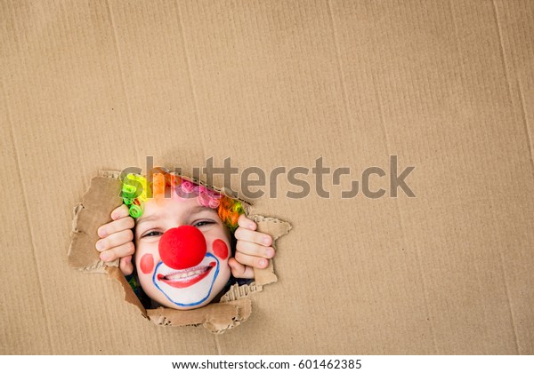 Think outside the box. Funny kid clown looking\
through hole on cardboard. Child playing at home. 1 April Fool\'s\
day concept. Copy space for your\
text.
