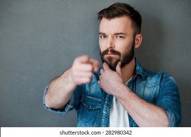 I think you must do it!Serious young man holding hand on chin and pointing you while standing against grey background
