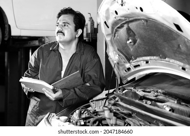 A Thinker Car Mechanic In Moustache Holding His Day Planner While Examining The Car. Car Specialist Is Standing Next To The Open Bonnet And Thinking. Repairman Wearing A Blue Mechanic's Uniform.