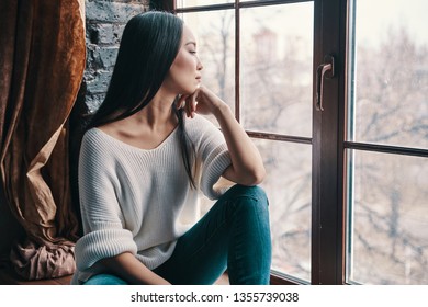 Thinking about.... Attractive young woman keeping hand on chin and looking away while sitting on the window sill at home