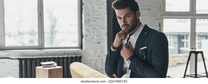 Thinking about... Handsome young man in full suit adjusting his shirt while leaning on the sofa - Powered by Shutterstock